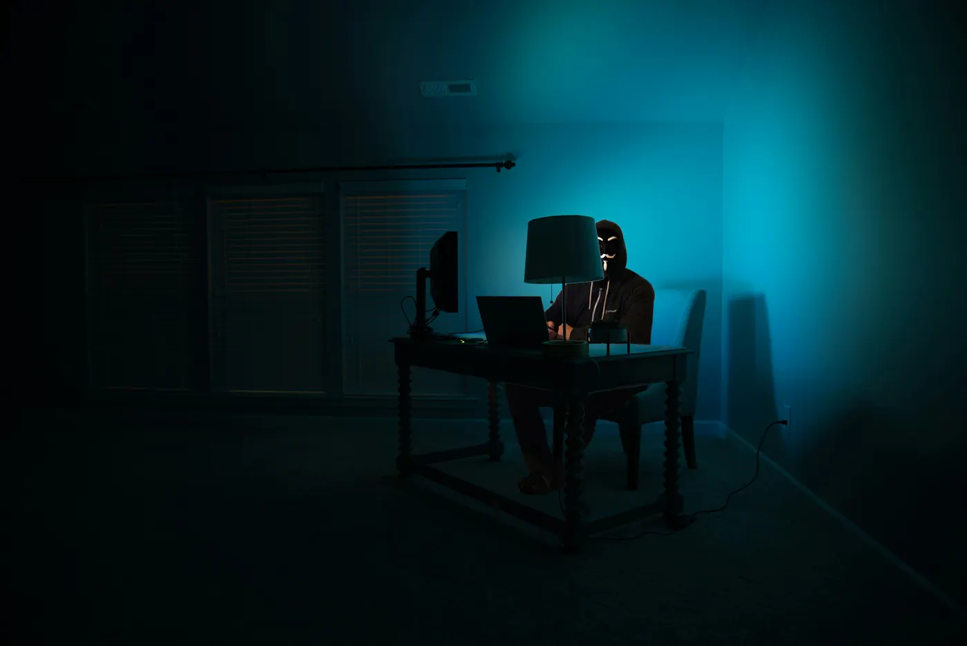 A hooded figure illuminated by cyan monitor light — representing the threat actor